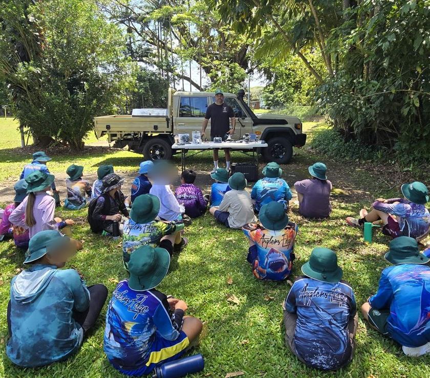 John giving a drone safety briefing beside the LandCruiser to a group of students sitting on the grass, seen from behind
