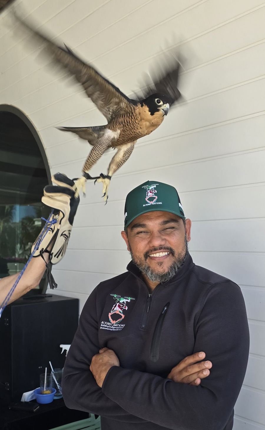 John Fejo in an AI First Nations cap and jacket, smiling, with a falcon taking off beside him